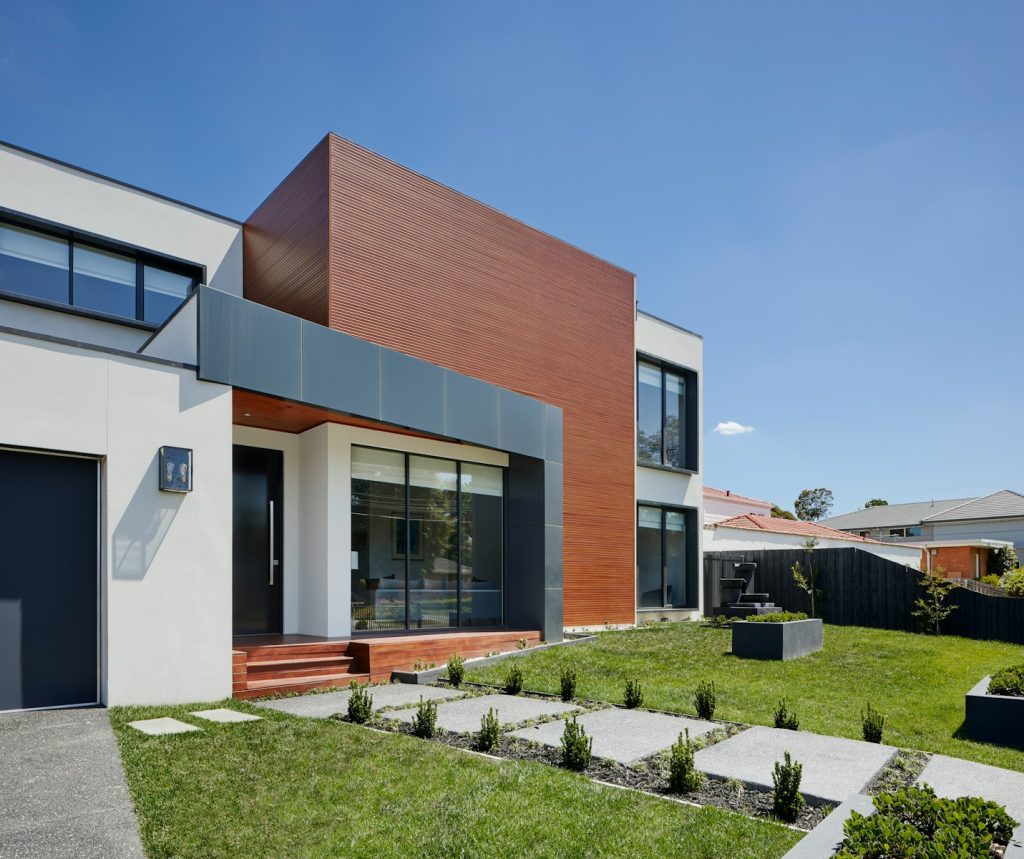Modern house exterior with sleek design, wooden paneling, large windows, and landscaped garden on a sunny day