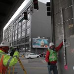 Surveyors measuring under an urban overpass, using equipment, wearing safety gear. City street in the background