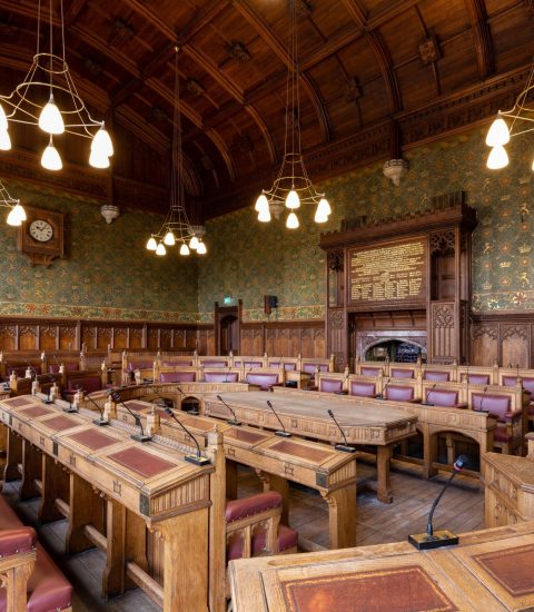 Ornate wood-panelled parliamentary chamber with green wallpaper, chandeliers, and microphones on wooden desks