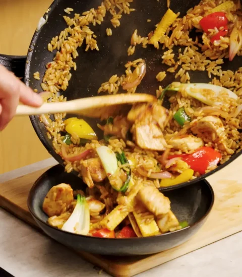 Chef serving vibrant stir-fried rice with vegetables and chicken from a wok into a black bowl on a wooden board