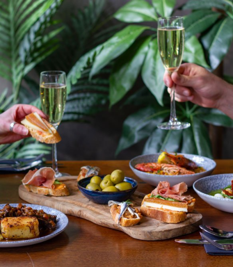 A vibrant dining scene with tapas and two people toasting with champagne, amidst lush green plants