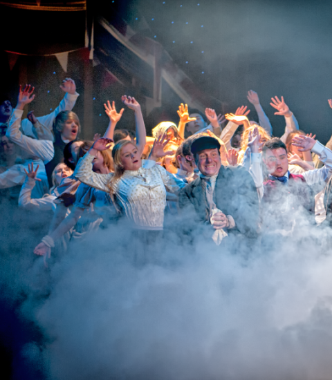 Theatrical performance with cast in dramatic smoke, actors expressing emotion in vibrant stage lighting