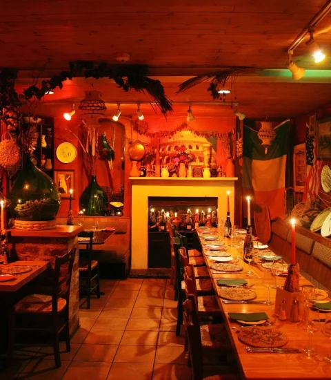 Cosy Italian-themed restaurant interior with warm lighting, long dining table, decorative wine displays, and rustic charm