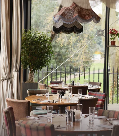 Cosy restaurant interior with set tables, colourful chairs, vintage lampshade, and garden view through floor-to-ceiling windows