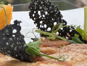 Gourmet dish with crispy black garnish, tender meat, and golden polenta on a white plate, garnished with fresh greens