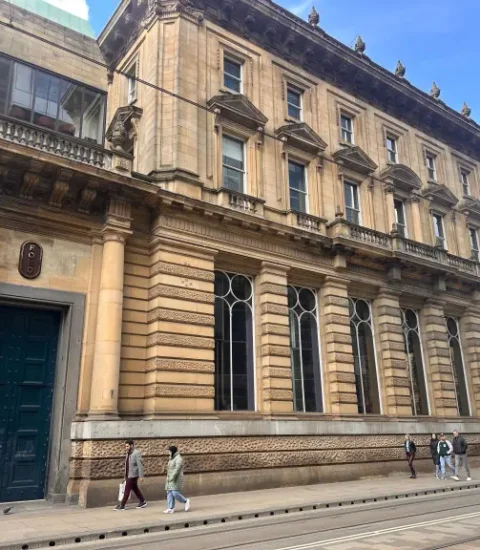 Historic stone building with large windows on a city street in Manchester, people walking by on a sunny day