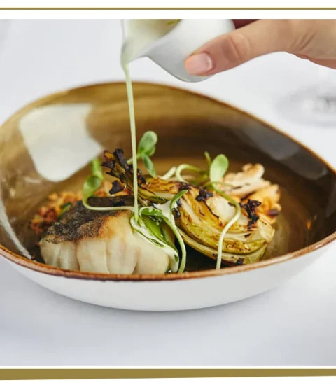 Hand pouring sauce over grilled fish and vegetables in elegant dish, gourmet presentation on white tablecloth