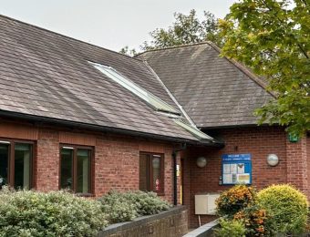Red-brick community centre with slate roof, surrounded by greenery and trees, under an overcast sky in England