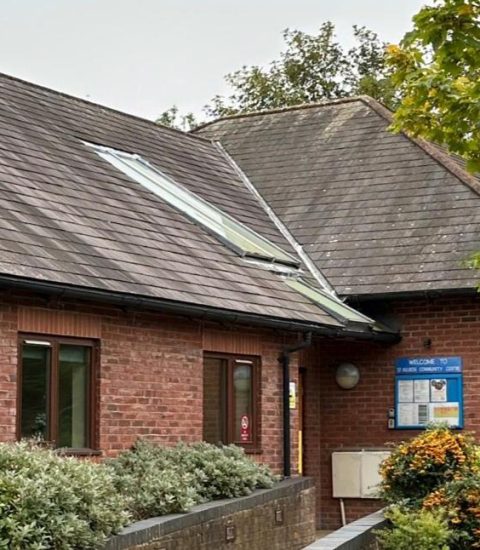 Red-brick community centre with slate roof, surrounded by greenery and trees, under an overcast sky in England