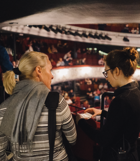 Two women talking in a theatre balcony with an audience seated below, creating an atmosphere of anticipation and excitement