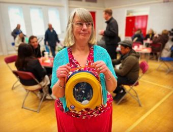 Woman holding a defibrillator in a community centre surrounded by people seated at tables, engaging in conversation