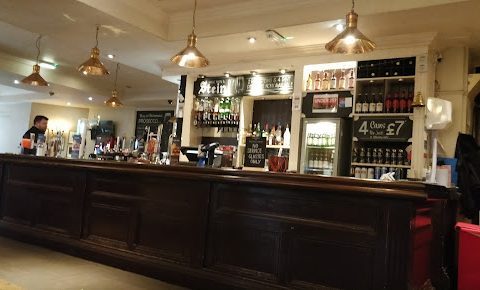 Cosy pub interior with a wooden bar, chalkboard menu, and hanging copper lights, offering drinks and snacks.