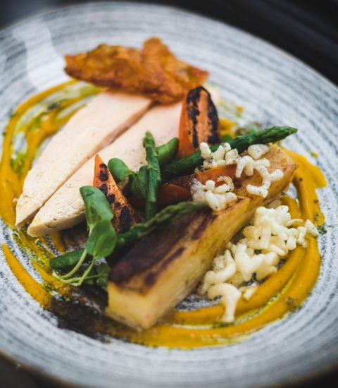 Gourmet dish featuring sliced chicken breast, roasted vegetables, asparagus and crispy garnish on a textured plate