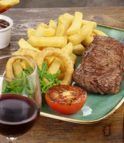Steak, chips, onion rings, and grilled tomato on a plate with red wine and sauce, perfect for a classic meal out