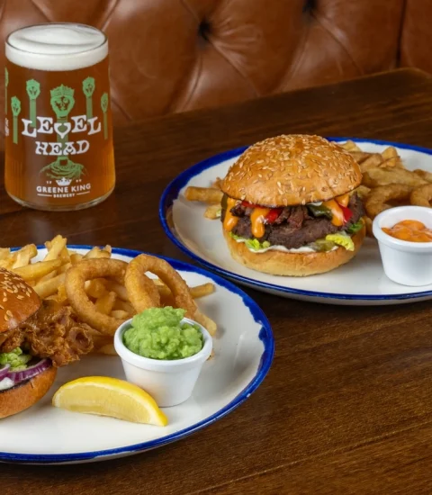 Two gourmet burgers with onion rings and chips, served with a pint of ale on a pub-style wooden table setting