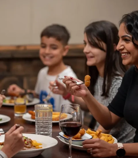 Group enjoying a meal at a restaurant, laughing and sharing food, with plates, glasses, and happy expressions visible