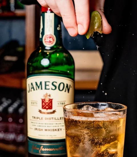 Hand squeezing lime into whiskey cocktail with Jameson bottle in background, creating a refreshing Irish drink scene