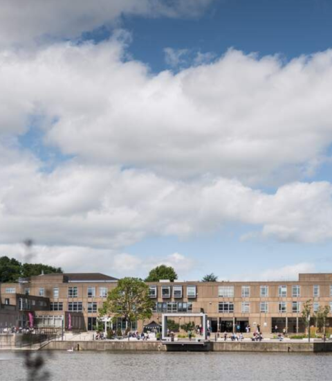 University campus with lake and modern buildings under a blue sky with clouds, featuring trees and a water fountain