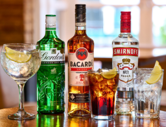 Selection of gin, rum, and vodka bottles with mixed drinks on a wooden table in a pub setting, featuring lemon slices