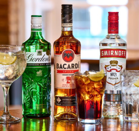 Selection of gin, rum, and vodka bottles with mixed drinks on a wooden table in a pub setting, featuring lemon slices