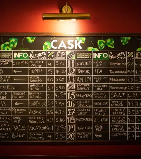 Pub chalkboard menu with various beers, breweries, and prices under a warm light in a cosy UK bar setting