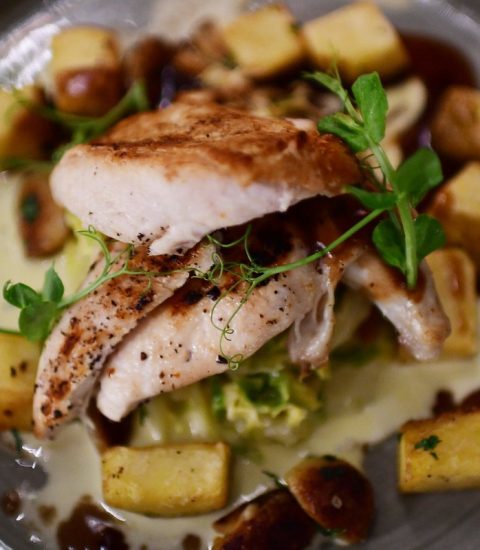 A gourmet dish featuring grilled chicken, roasted potatoes, creamy sauce, and pea shoots on a grey ceramic plate