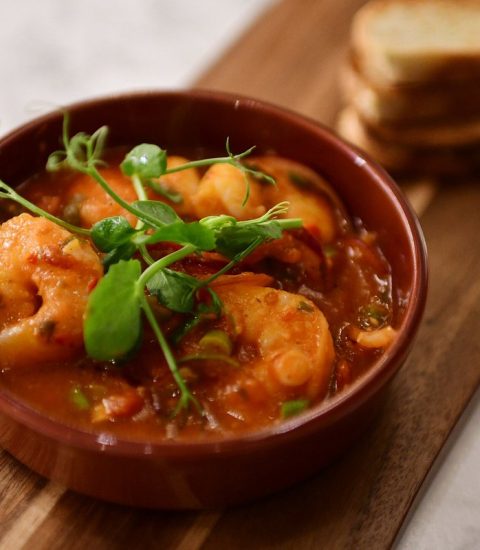 Prawns in rich tomato sauce garnished with greens, served in a bowl alongside toasted bread slices on a wooden board