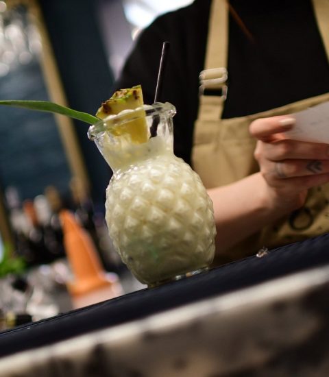 Tropical pineapple-themed cocktail on bar counter with bartender holding a receipt in the background, close-up shot