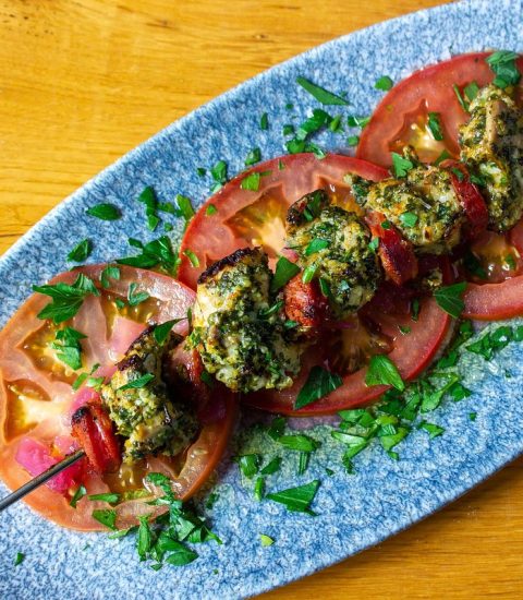 Grilled kebabs with herbs on sliced tomatoes, served on a blue plate, garnished with fresh parsley leaves