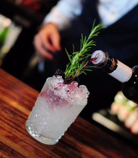 Bartender pouring red wine into a cocktail glass with ice and rosemary garnish on a wooden bar counter