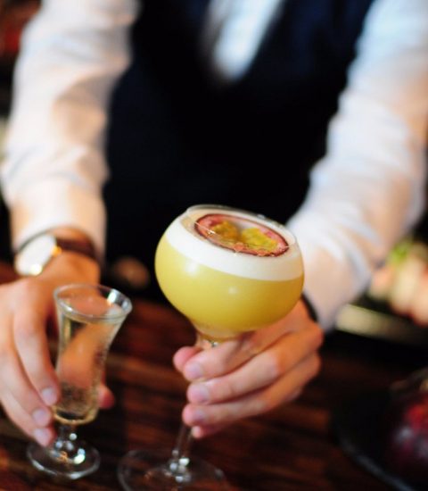 Bartender serving a yellow passionfruit cocktail with a shot on the side at a wooden bar counter