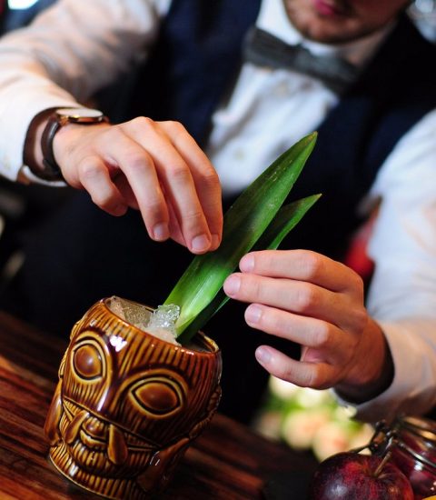 Bartender crafting a tropical cocktail in a tiki mug, garnished with fresh leaves and ice cubes at a stylish bar counter