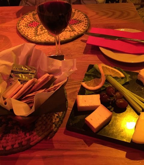 Cheese platter with assorted cheeses, grapes, celery, and orange slices, accompanied by breadsticks and red wine on a table