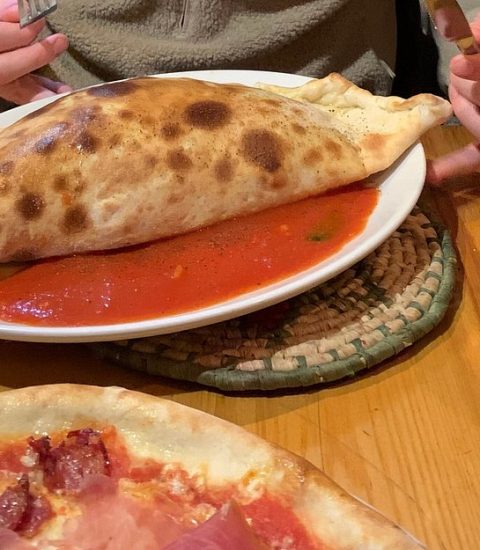 Calzone with tomato sauce on a table alongside a pizza, glass of water, and a drink with a straw, ready to enjoy