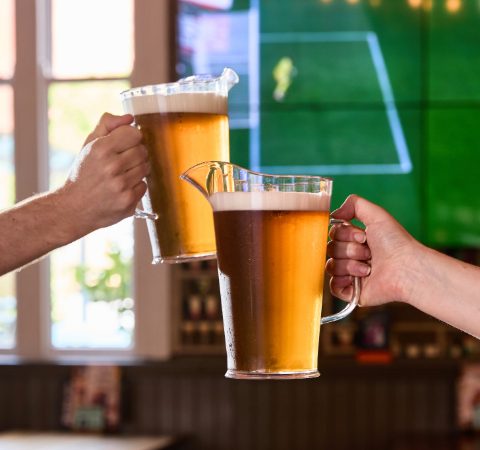 Two people clinking beer pitchers in a pub with a football match on TV in the background, celebrating sports victory