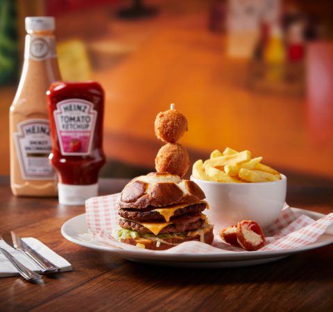 Gourmet burger with cheese, chips, and sauces on a wooden table, accompanied by ketchup and smoky BBQ sauce bottles