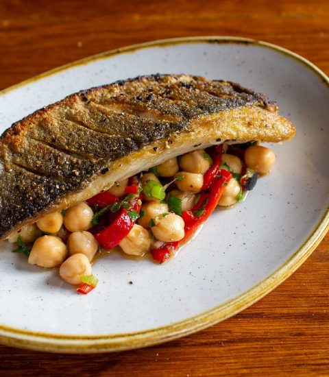 Grilled fish with crispy skin served on a bed of chickpeas and roasted red peppers on a rustic ceramic plate.