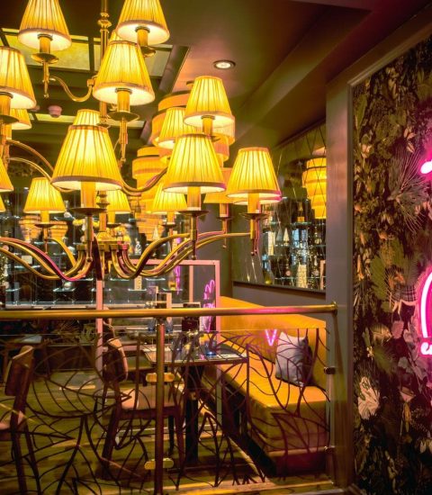 Chic restaurant interior with elegant chandeliers and neon enjoy it all sign on floral wall, warm ambient lighting