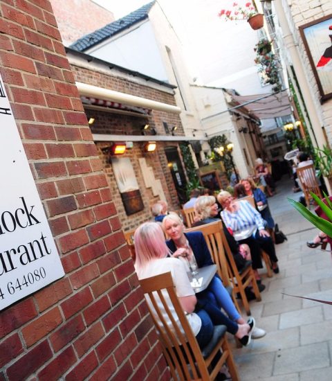 Outdoor dining at a lively restaurant with patrons enjoying meals, framed by a large clock-themed sign on a brick wall