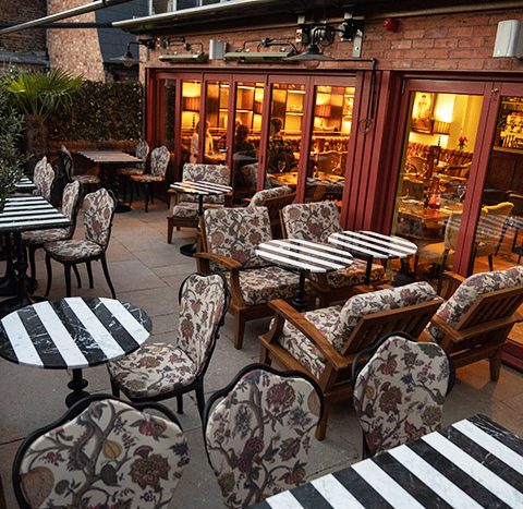 Charming outdoor café with striped tables and floral seating, warmly lit interior visible through glass doors