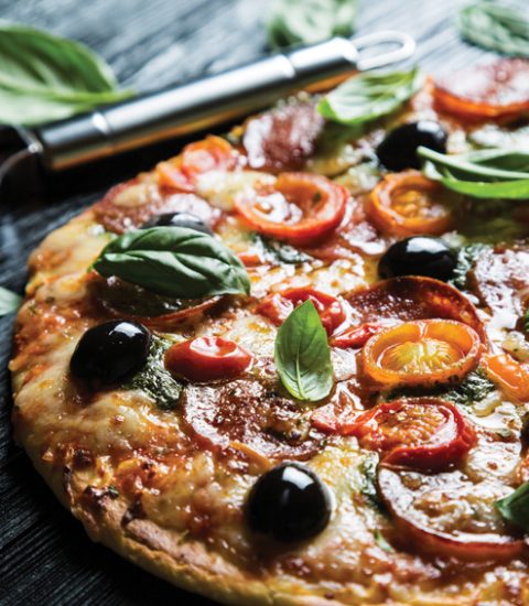 Delicious pepperoni pizza with olives and fresh basil on wooden table with cutter visible