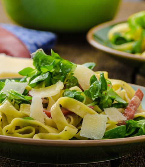 Fresh tagliatelle pasta with rocket, parmesan, and prosciutto served on a green plate, garnished with lemon wedges.