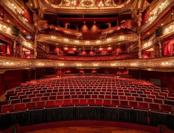 Ornate interior of a grand theatre with red velvet seats, golden decor, and detailed balconies, captured from stage view