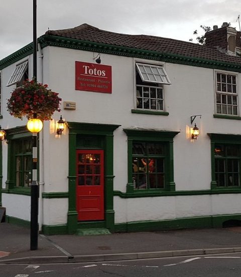 Exterior of a traditional British pub with green accents, a red door, hanging flowers, and classic street lamps.