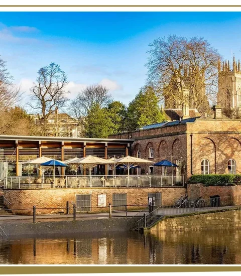 Historic riverside building with outdoor seating, surrounded by trees and blue sky, ideal for dining or leisure in the UK