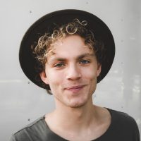 Young man smiling, wearing a black hat and a grey top, curly hair, and standing against a neutral background